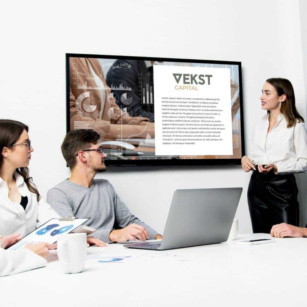 A woman stands and presents a slide for VEKST Capital to three colleagues in a modern office, highlighting their custom logo design services as they work with laptops, papers, and coffee cups.