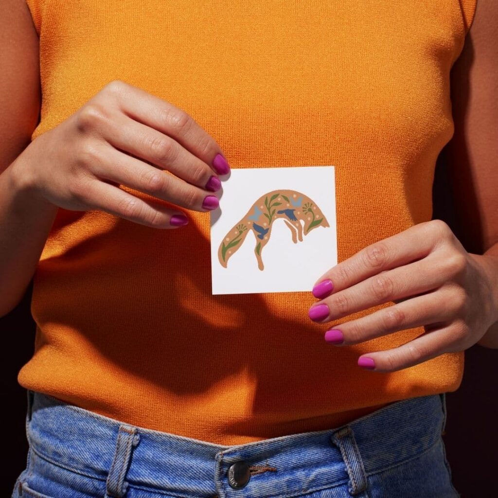 A person in an orange top and blue jeans holds a square card with an illustrated fox and decorative foliage, showcasing custom logo design. The persons nails are painted purple.