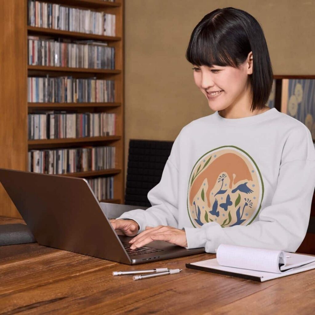 A woman with short dark hair smiles while working on a laptop at a wooden table. She wears a light-colored sweatshirt with an abstract animal design, perhaps brainstorming ideas for custom website design. Nearby are a notepad, pen, and shelves filled with CDs.