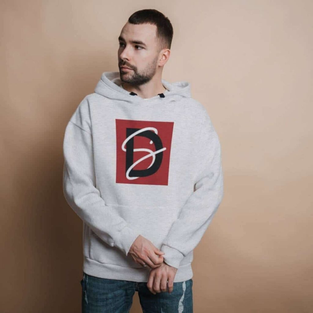 A man wearing a light gray hoodie featuring a custom logo design in red and black stands against a plain beige background, looking to his left with his hands loosely clasped in front.