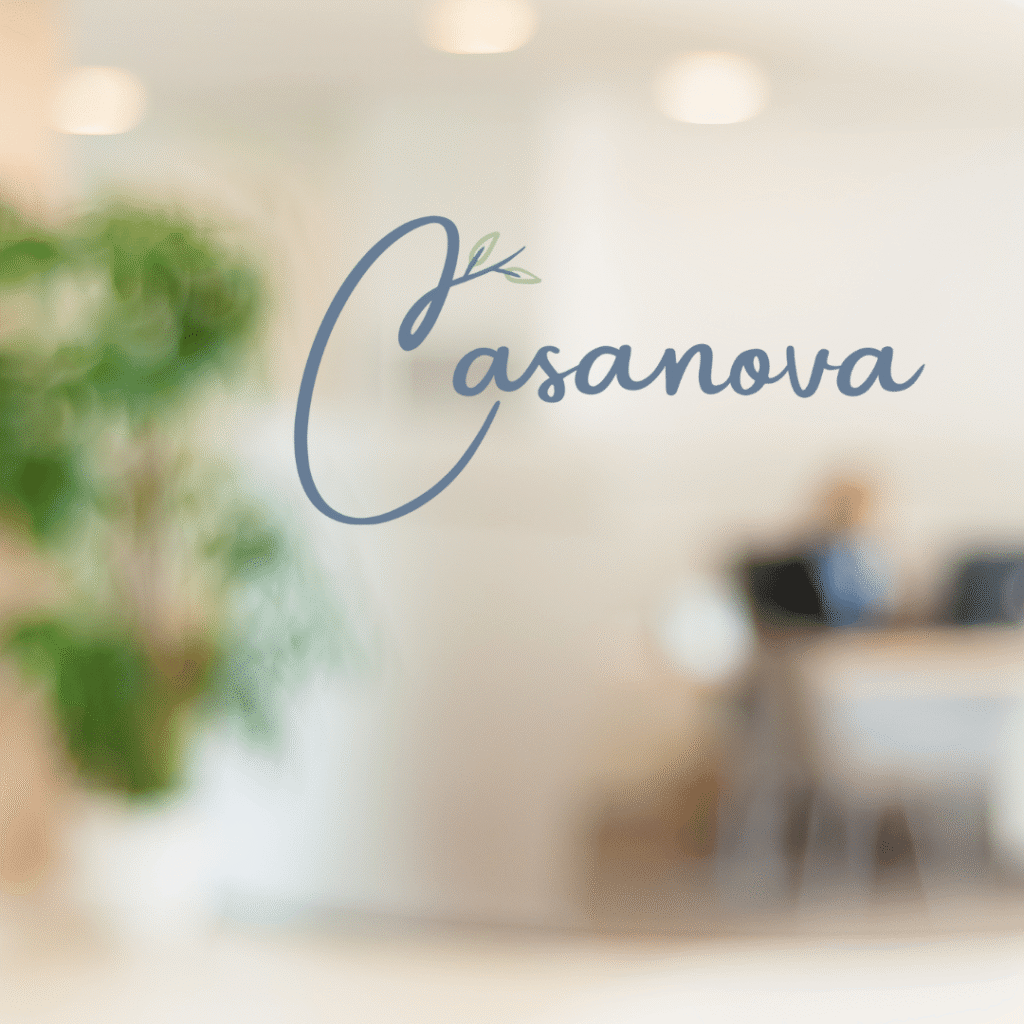 The word Casanova in elegant script is displayed on a glass wall, showcasing Moonlit Medias logo design services; a leafy accent graces the letter C, with a blurred office setting and green plant in the background.
