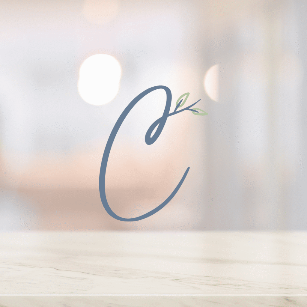 A large, stylized letter C with a small green leafy branch emerging from its top curve, set against a blurred background and light marble surface—perfect for those seeking unique logo design services from Moonlit Media.
