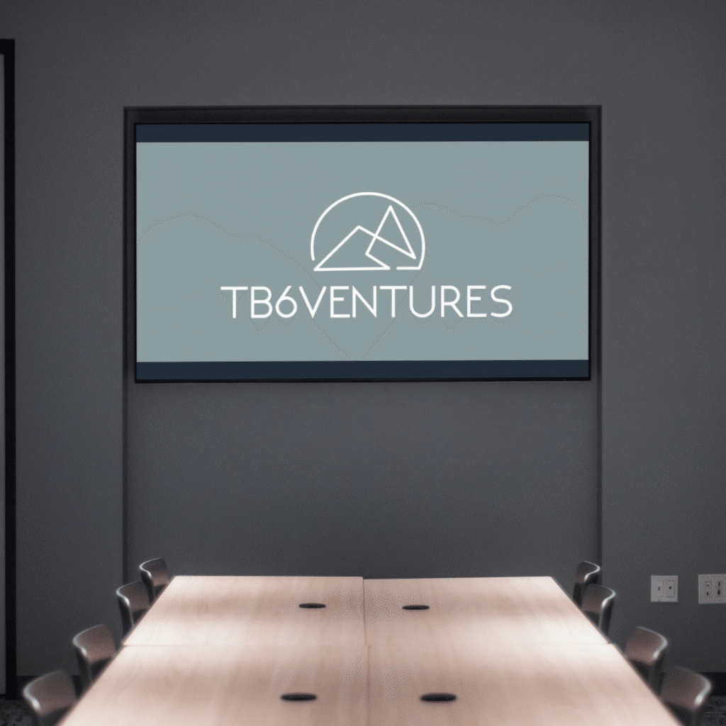 A conference room with a wooden table and chairs, featuring a large screen on the wall displaying the TB6Ventures logo with a geometric mountain design—an example of custom logo design by Moonlit Media.
