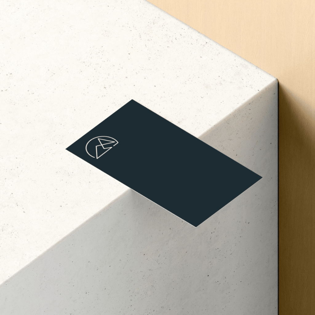 A dark business card with a simple geometric logo in the corner rests partially over the edge of a light-colored, textured stone surface—a sleek display for Moonlit Media’s professional logo design services.