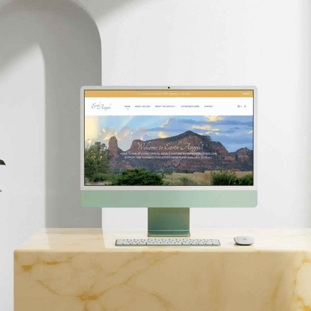 A desktop computer on a marble desk displays a custom website design with a scenic landscape of red rock formations and greenery under a cloudy sky. A white wireless keyboard and mouse are placed on the desk.