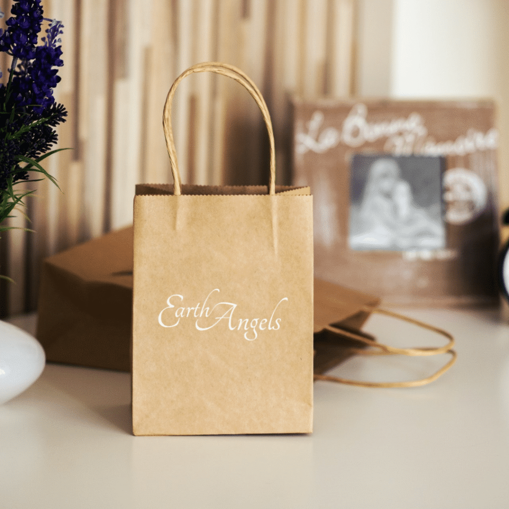 A small brown paper gift bag with a custom logo design reading Earth Angels printed in white sits on a white surface next to a purple flower arrangement and another paper bag in the background.