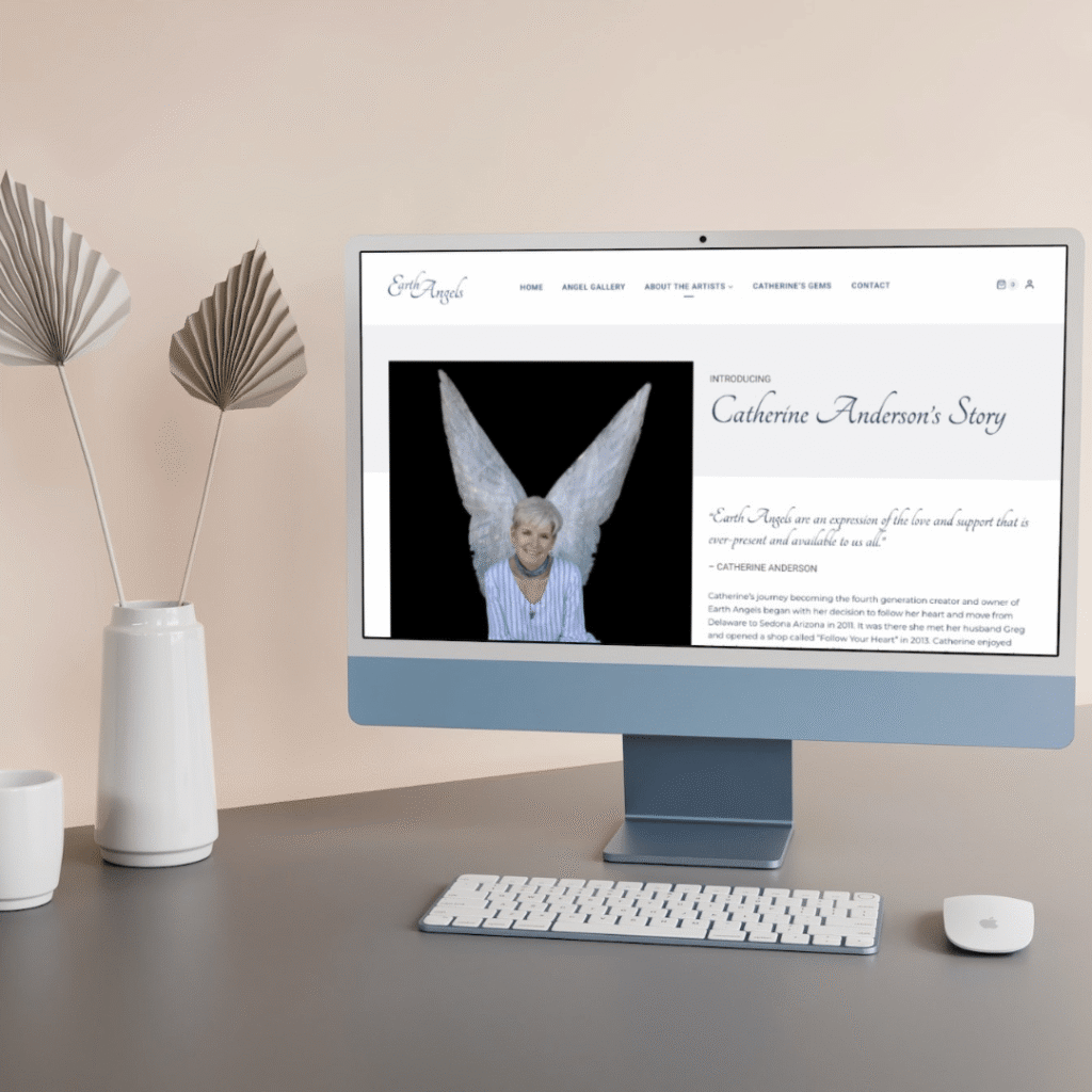A computer monitor on a desk displays a custom website design by Moonlit Media, featuring Catherine Anderson’s Story with an image of an older woman smiling in angel wings. The desk also includes a keyboard, mouse, vase, and decorative fan-shaped objects.