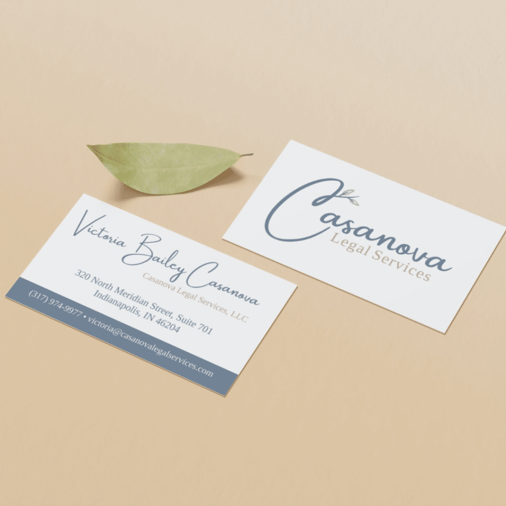 Two business cards for Casanova Legal Services, designed by Moonlit Media, rest on a beige surface beside a green leaf. One card highlights their custom logo design; the other features Victoria Bailey Casanova’s contact information.