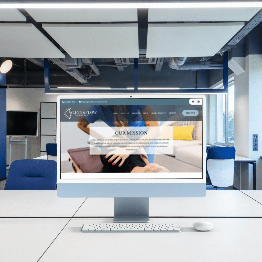 A modern office with a large computer monitor on a desk, showcasing the Align and Flow Chiropractic website—crafted by Moonlit Media’s custom website design. The space features blue chairs, white tables, and ceiling panels.