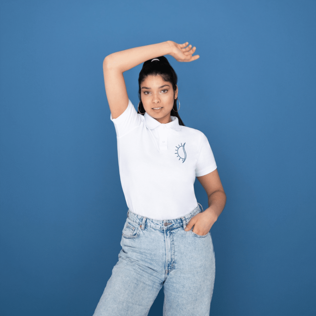 A woman poses confidently against a blue background, wearing a white polo shirt with an eye graphic—perfectly embodying Moonlit Media’s modern touch, much like their custom website design. She raises one arm and looks directly at the camera.