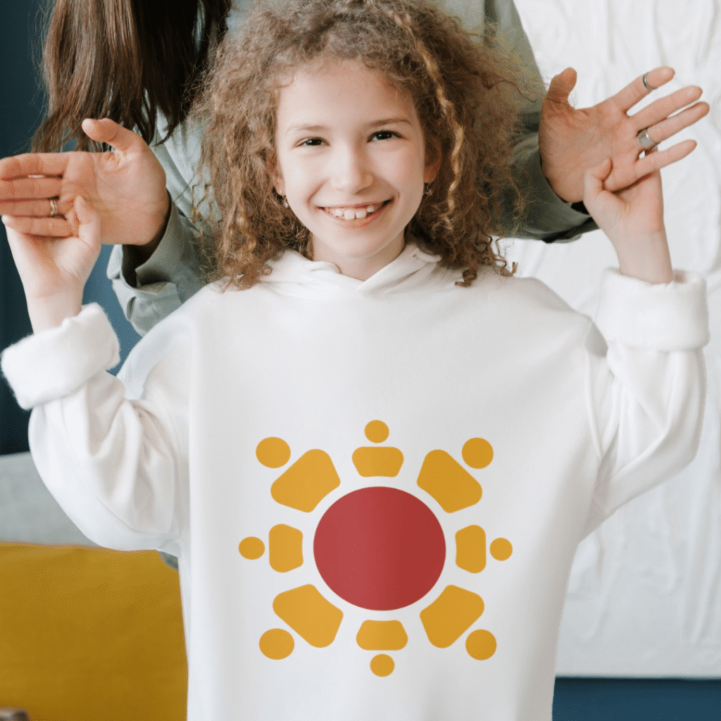 A smiling child in a white hoodie with a red and yellow abstract sun—showcasing a custom logo design—stands indoors, raising both hands, while an adult behind them playfully holds up their hands as well.