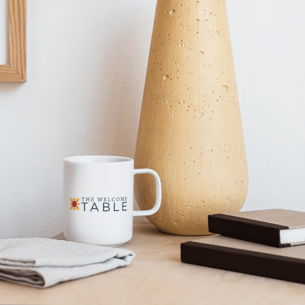 A white mug with THE WELCOME TABLE printed on it, designed by Moonlit Media’s logo design services, sits on a wooden surface next to a tall beige vase, a folded cloth, and two closed notebooks against a plain white wall.