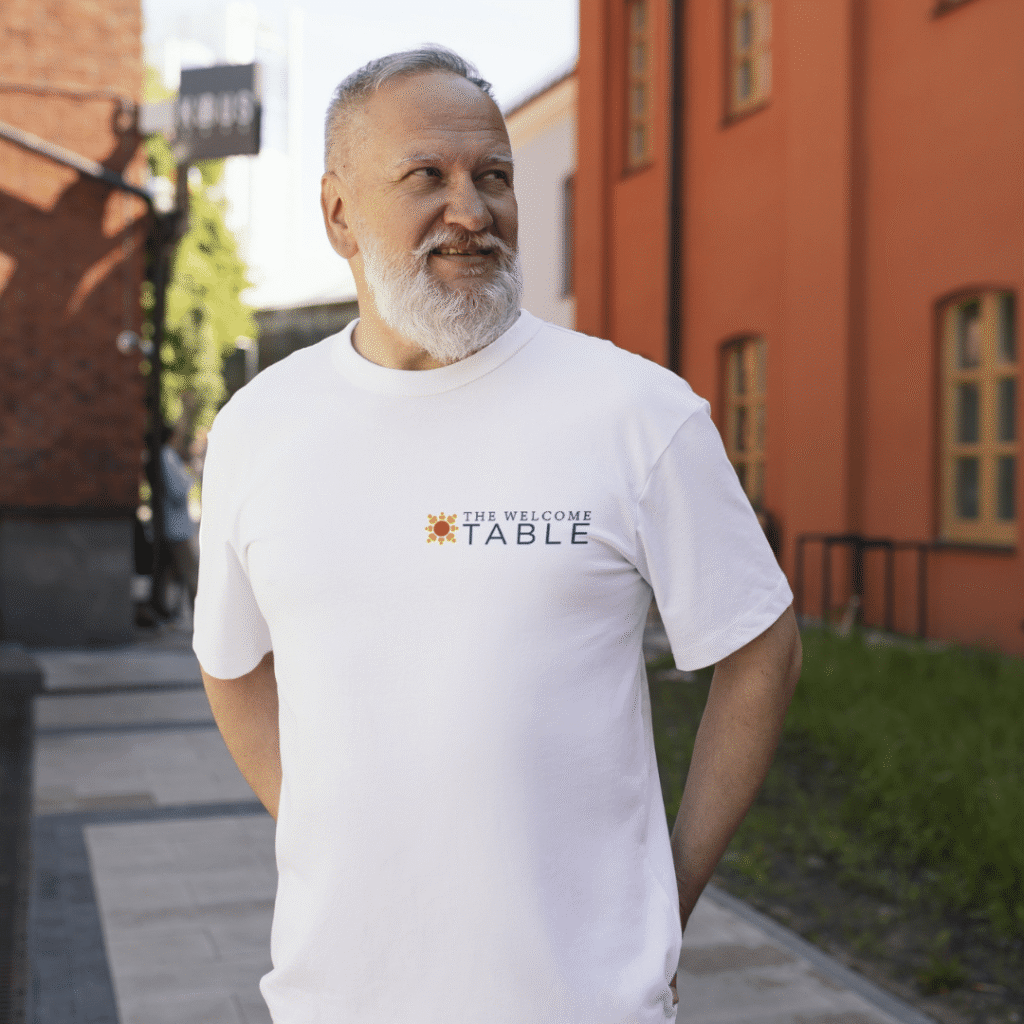 An older man with a white beard stands outdoors, smiling and wearing a white t-shirt that reads THE WELCOME TABLE with a sun icon—behind him are red and gray buildings and a sidewalk, reflecting the charm of thoughtful logo design services.