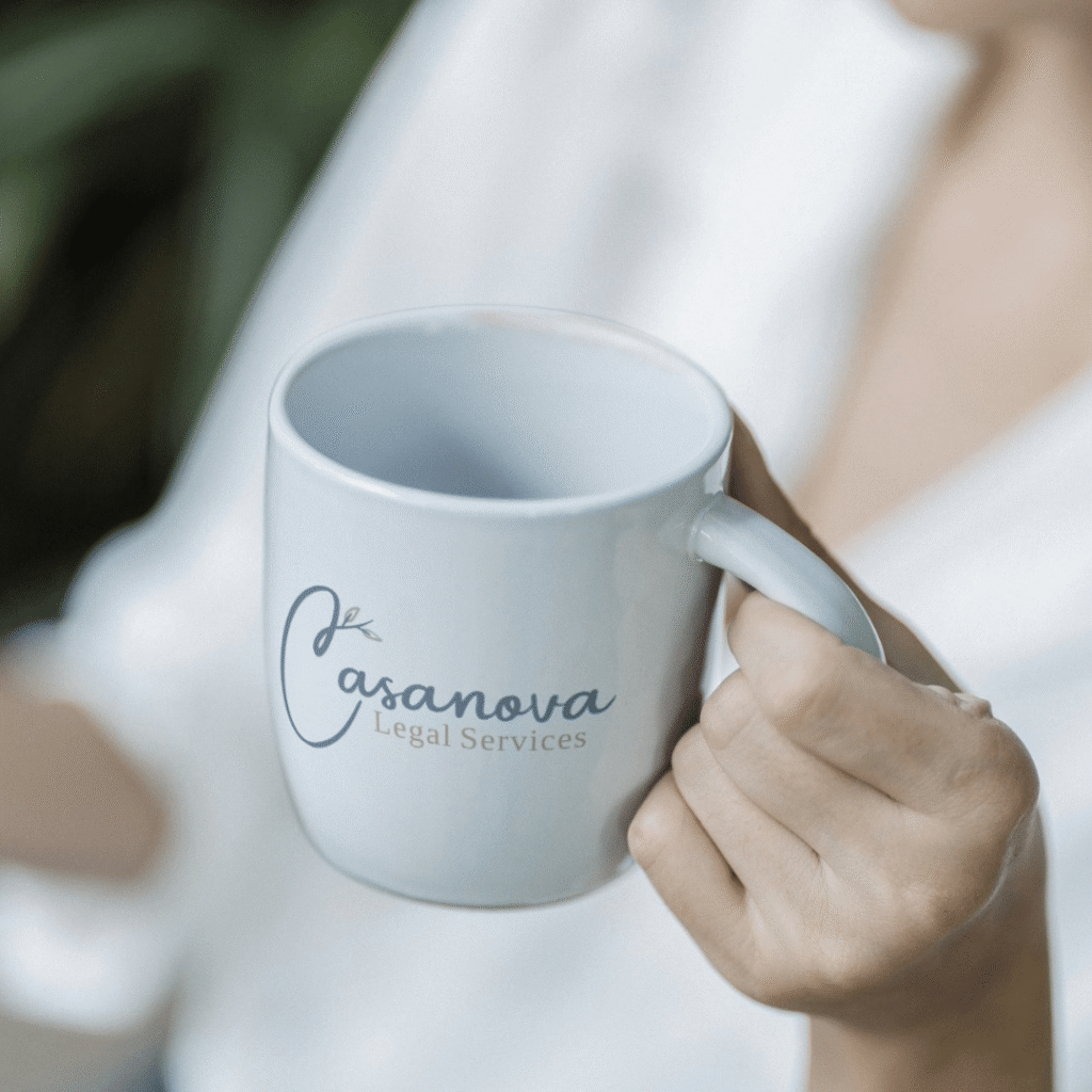 A person holds a white mug featuring Casanova Legal Services in blue script, showcasing a custom logo design by Moonlit Media. The background is blurred, directing all attention to the branded mug and the hand holding it.