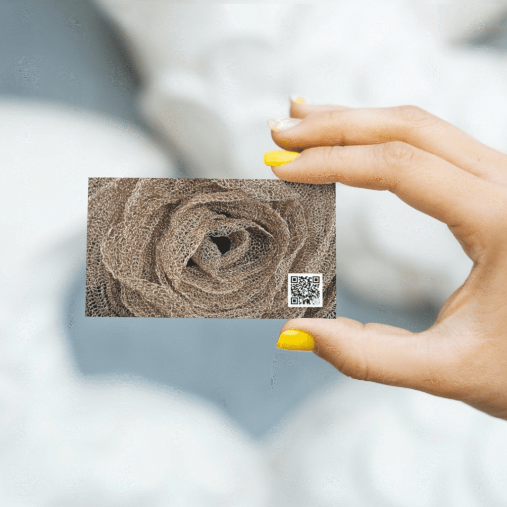 A hand with yellow-painted nails holds a business card for Moonlit Media, featuring a close-up textured rose and a small QR code. The softly blurred background highlights their web design services.