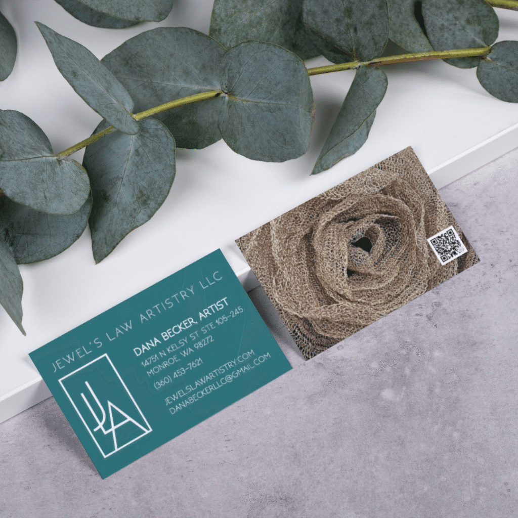 Two business cards rest on a white surface next to eucalyptus leaves. One card shows contact details for Dana Becker, artist at Jewels Law Artistry LLC; the other features a burlap rose, QR code, and highlights custom website design by Moonlit Media.