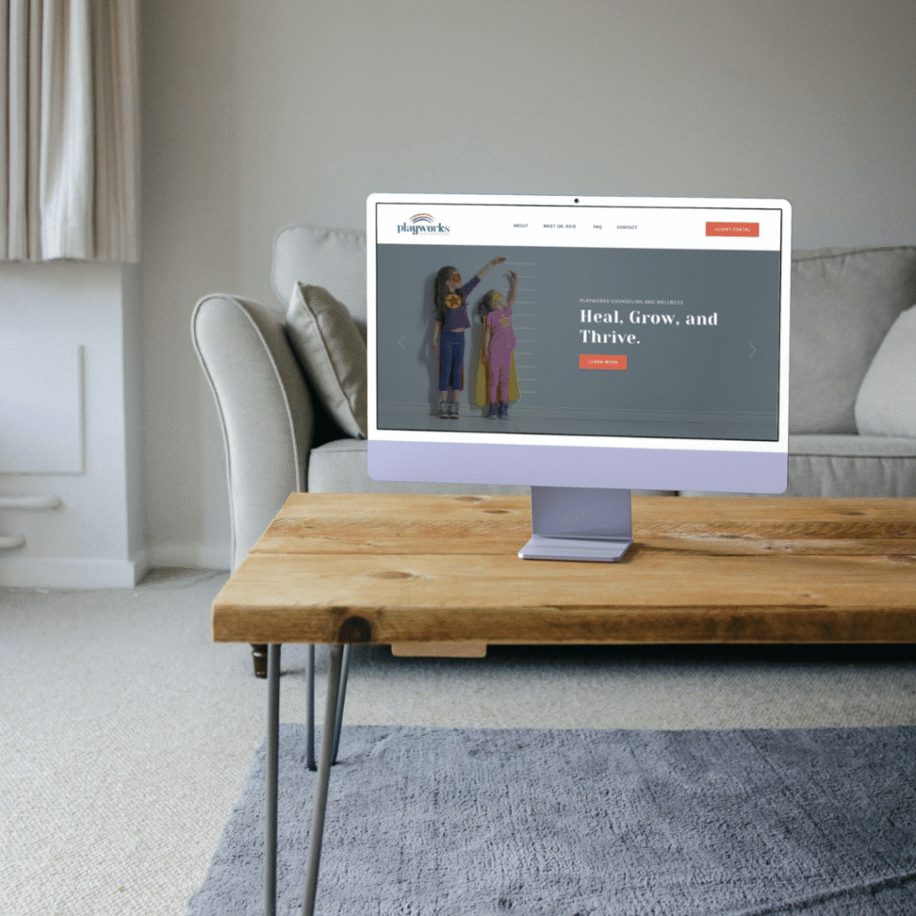 A computer monitor on a wooden coffee table displays a website with an image of two children and the words “Heal, Grow, and Thrive,” showcasing the work of a skilled web designer. The scene is set in a modern living room with a beige couch and carpet.