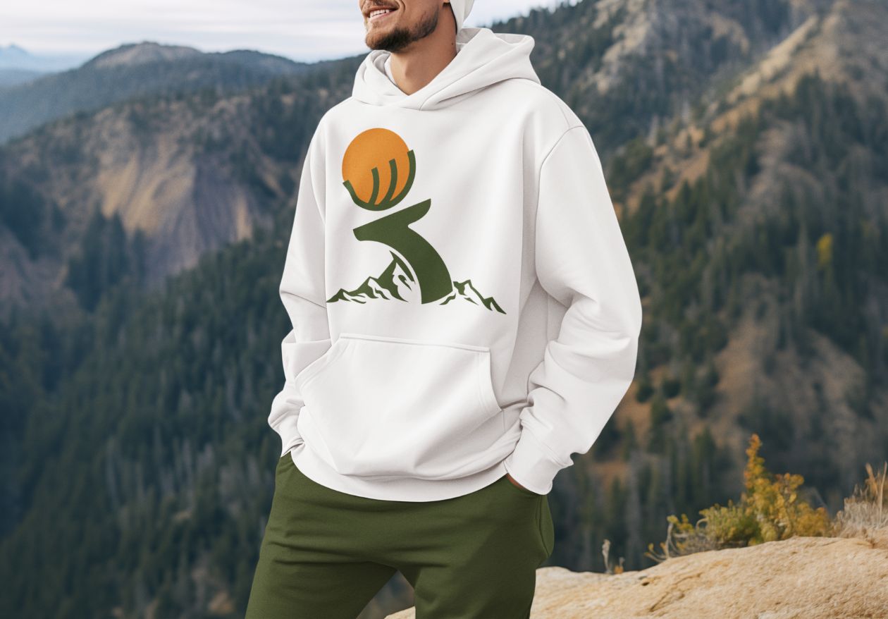 A person wearing a white hoodie with a graphic of a stylized sun, hand, and mountains stands outdoors with forested mountains in the background, representing the creativity found in moonlit media's logo design services.