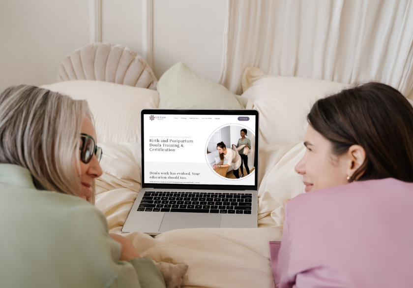 website design trends to watch and how to choose two women discussing a website. Two women lie on a bed facing a laptop, which displays a website about birth and postpartum doula training and certification, showcasing 2026 web design trends. The room features light bedding and a scalloped pillow in the background.
