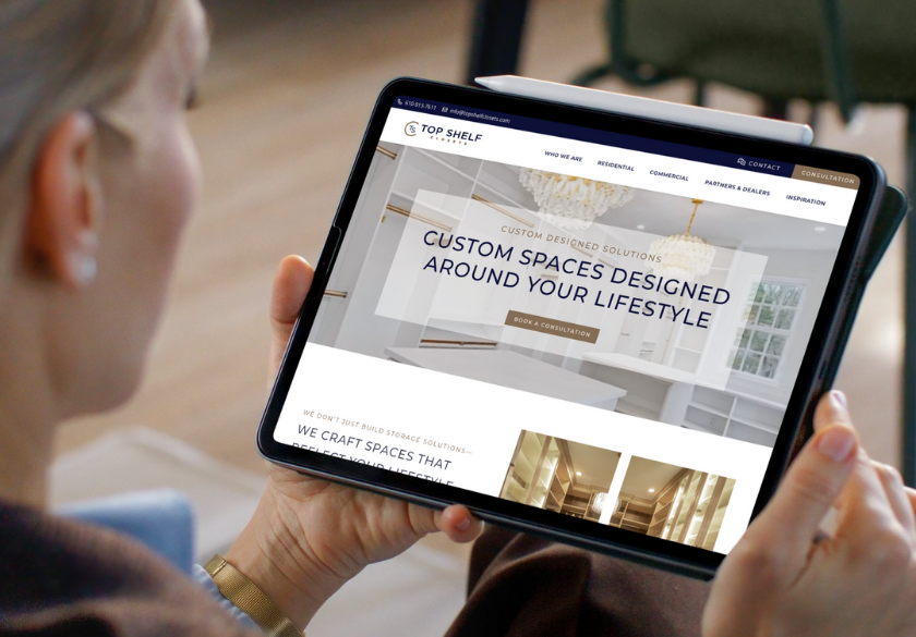 A person holds a tablet displaying the Top Shelf Design website, highlighting custom spaces and an image of a modern kitchen—showcasing emerging web design 2026 trends with sleek pendant lights and white cabinetry.