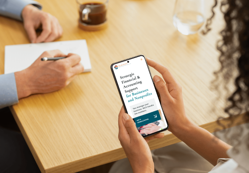 A person holds a smartphone displaying a financial and accounting support website, while another takes notes at a wooden table. This setup helps users brief a designer for best results in managing finances efficiently.