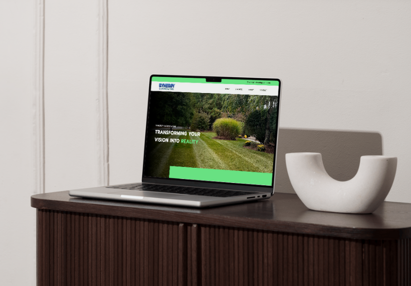 A laptop on a wooden cabinet displays a landscaping website, perfect for those looking to brief a designer for the best design results. A modern, white decorative object sits beside the laptop against a plain wall.