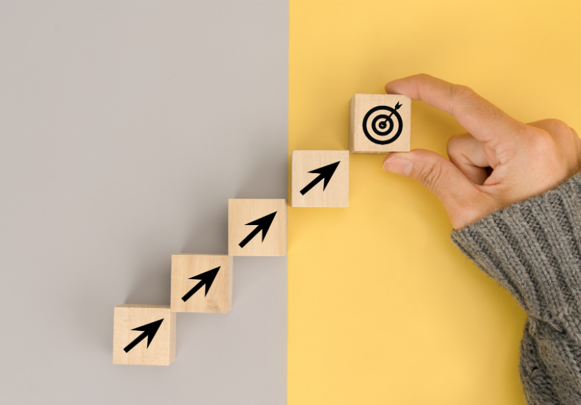 A hand places a wooden block with a target icon atop a staircase of blocks with arrows, symbolizing goal achievement and how to brief a designer for the best design results, against a gray and yellow background.