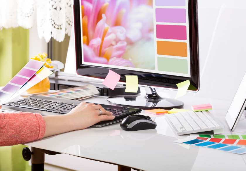 A person works at a desk with a computer displaying colorful swatches. Color samples, sticky notes, and a keyboard are on the desk, suggesting they’re preparing a designer brief to achieve the best results in their creative work.