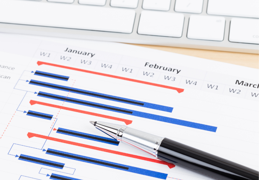 A close-up of a printed Gantt chart with red and blue bars, a black pen resting on it, and part of a white computer keyboard in the background—perfect for use in designer briefing sessions to achieve the best results.