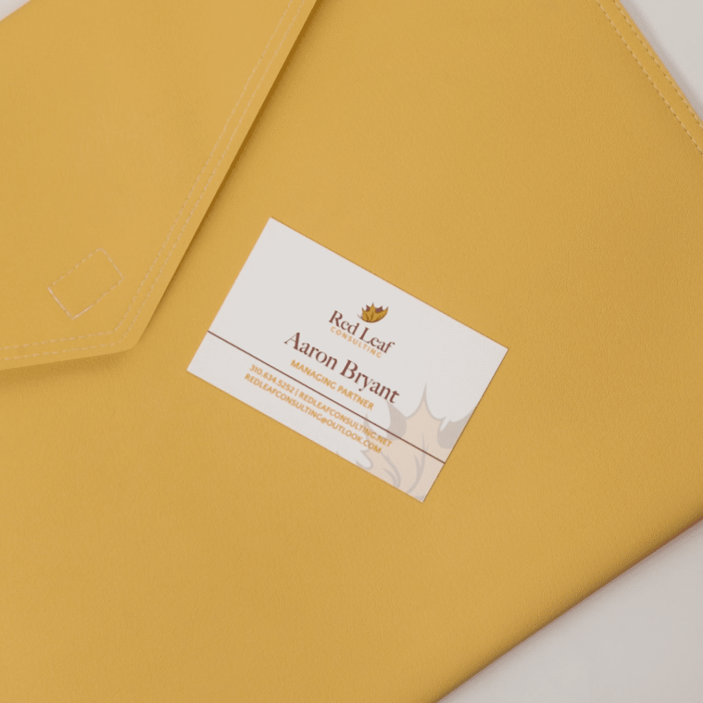 A business card for Aaron Bryant of Red Leaf is placed on a yellow leather folder. The card features a custom logo design by Moonlit Media, contact details, and a subtle leaf design in the background.