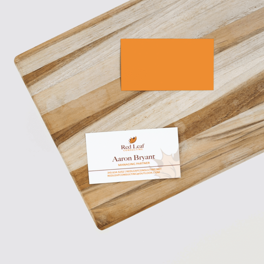 Two business cards on a light wooden board: one is blank and orange, the other is white with orange and brown text for Red Leaf Consulting—featuring Aaron Bryant, Managing Partner. Printed by Moonlit Media, known for custom website design and web design services.