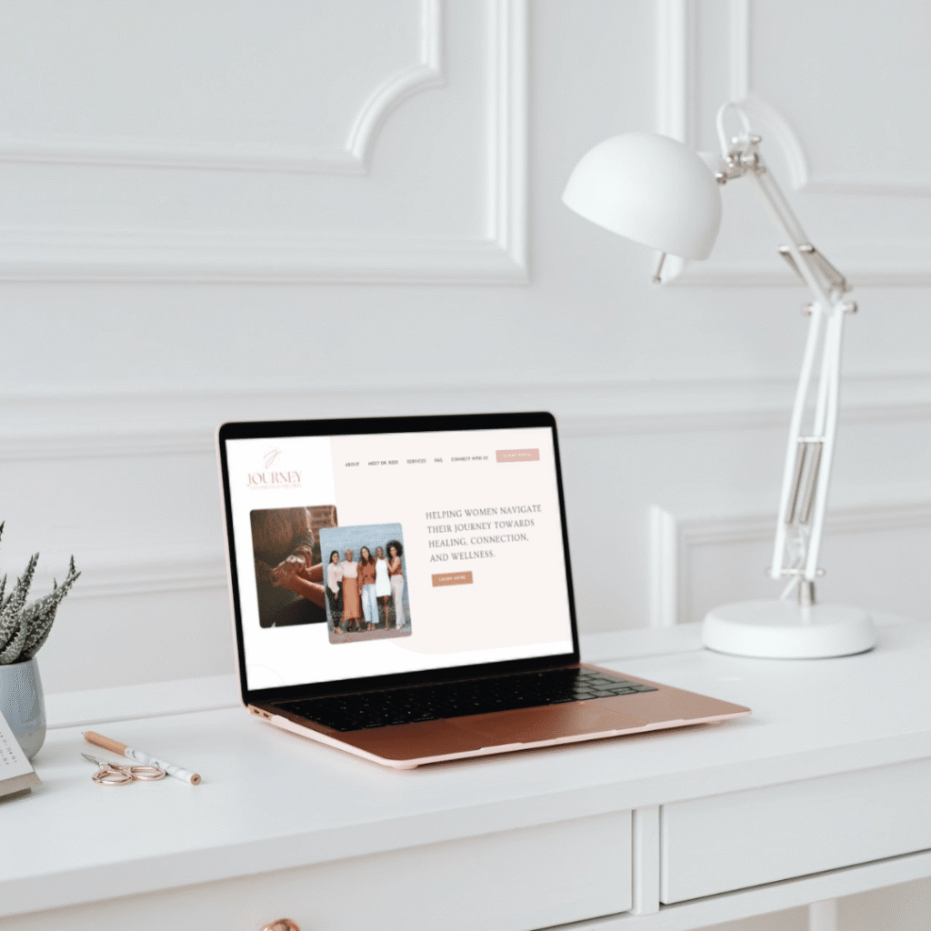 214 | Moonlit Media LLC A rose-gold laptop on a white desk displays a website titled Journey with photos of women. The desk also holds a lamp, a plant in a pot, and stationery—an inspiring scene for moonlit media or logo design services against a white-paneled wall.