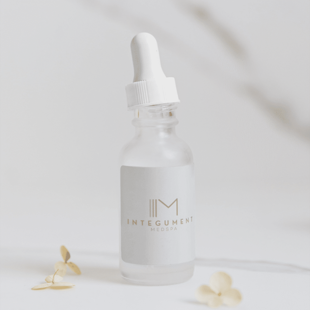 A frosted glass dropper bottle labeled INTEGUMENT MEDSPA sits on a white surface with beige flower petals nearby, set against a minimal, light background—perfect for showcasing logo design services from Moonlit Media.
