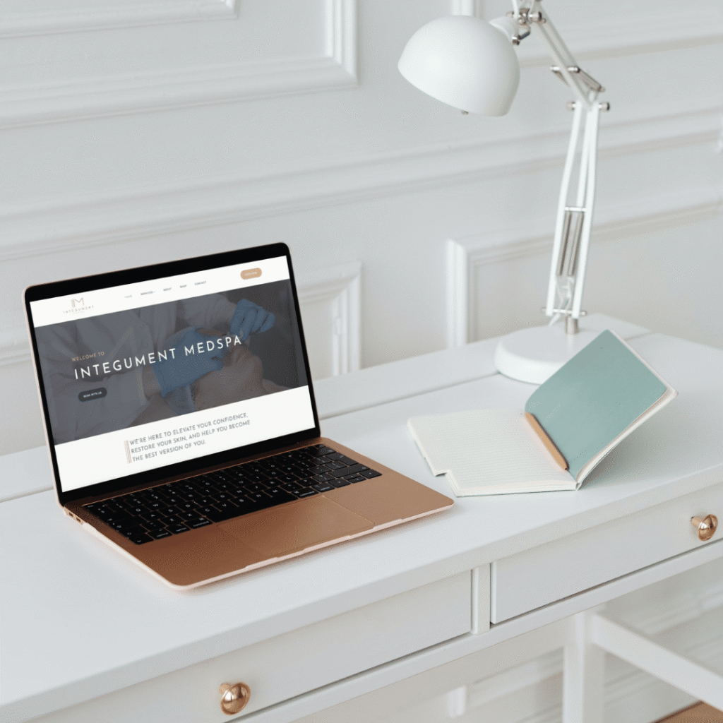 A gold laptop displaying a medspa website by Moonlit Media sits on a white desk next to an open notebook and a white desk lamp in a bright, minimalist room.