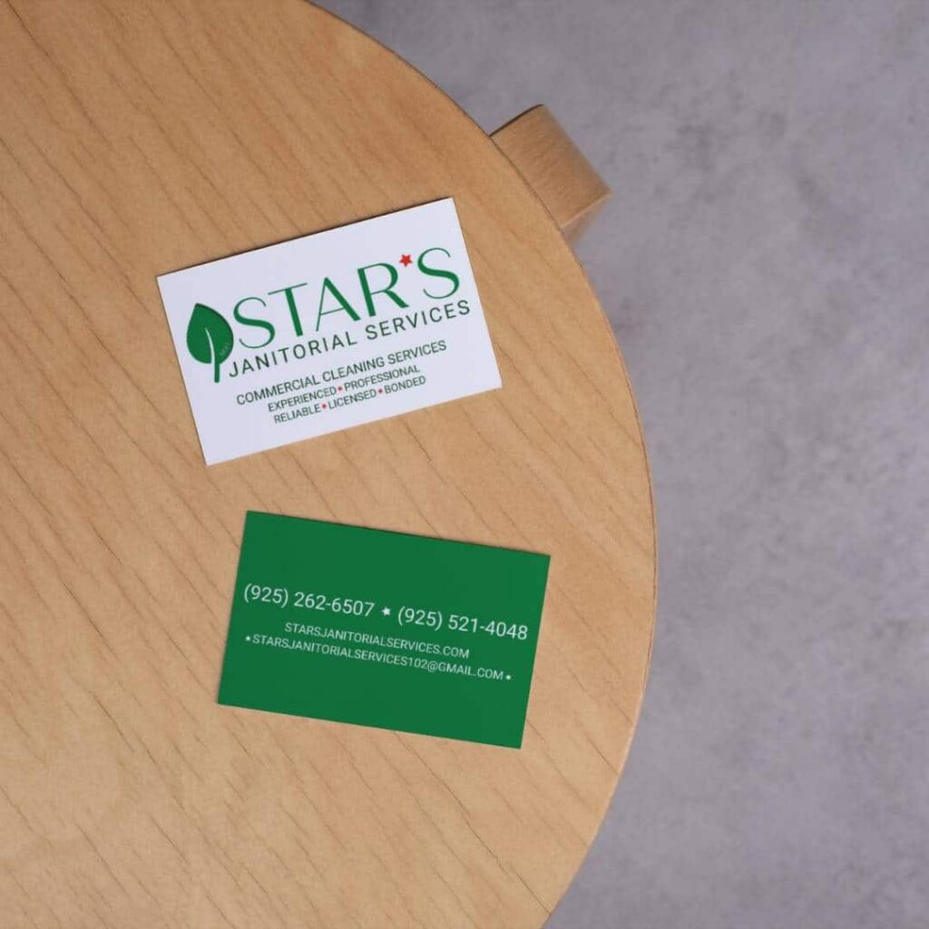 Two business cards for Stars Janitorial Services rest on a light wooden surface; alongside contact information, one card highlights their custom website design offerings in green and red text, while the other features white text on a green background.