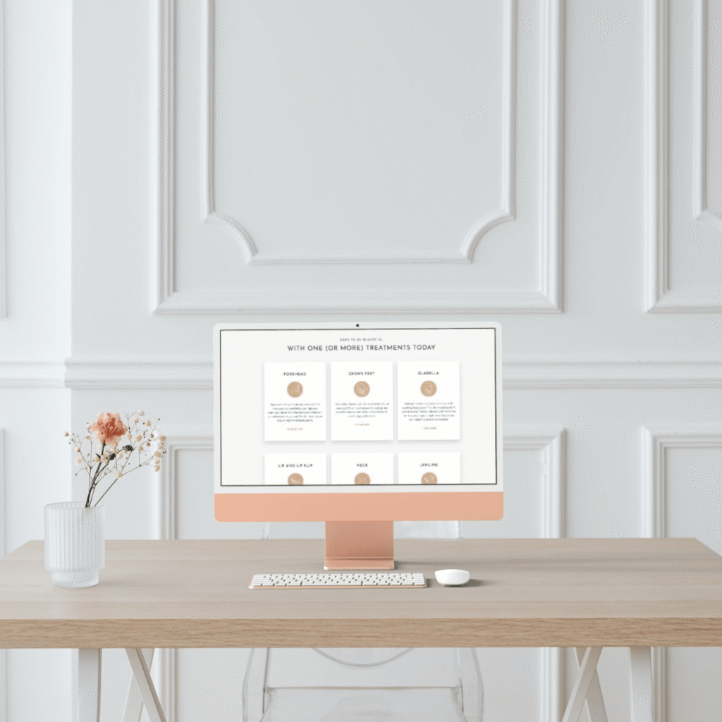 A minimalist workspace with a light wood desk, clear chair, vase of dried flowers, and a peach-colored desktop computer displaying custom logo design by Moonlit Media, set against a white paneled wall.