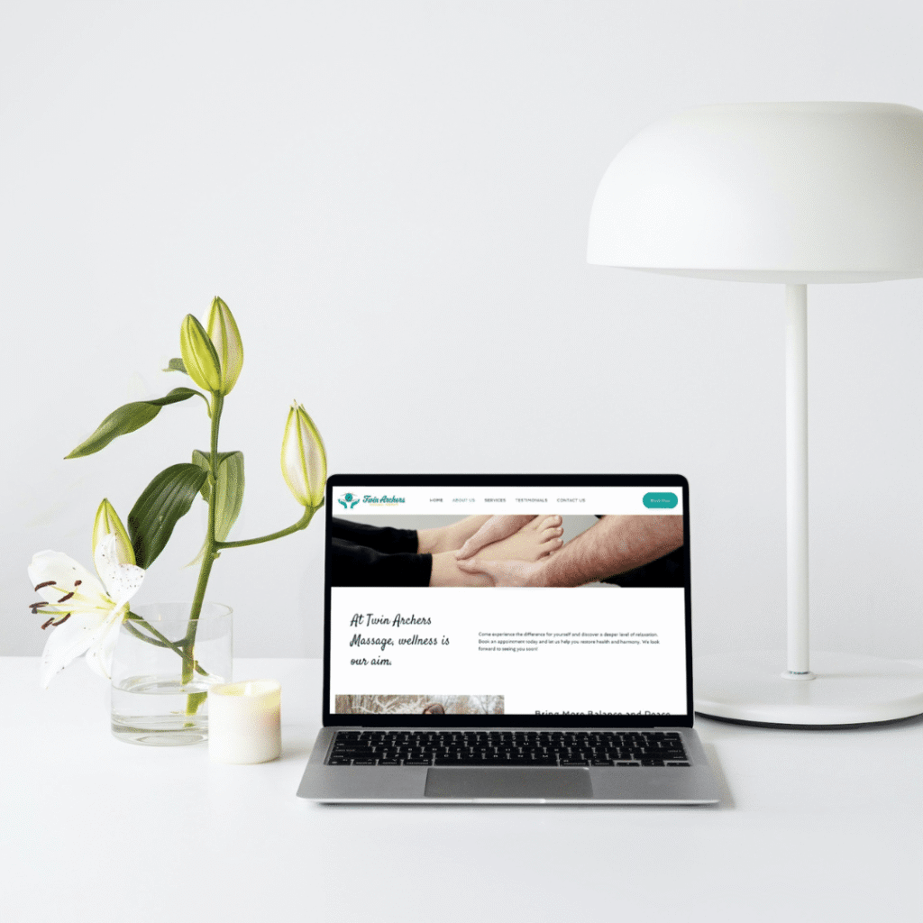 A laptop displaying a wellness website sits on a white desk beside a white lamp, a glass vase with white lilies, and a lit candle—perfect inspiration for web design services.