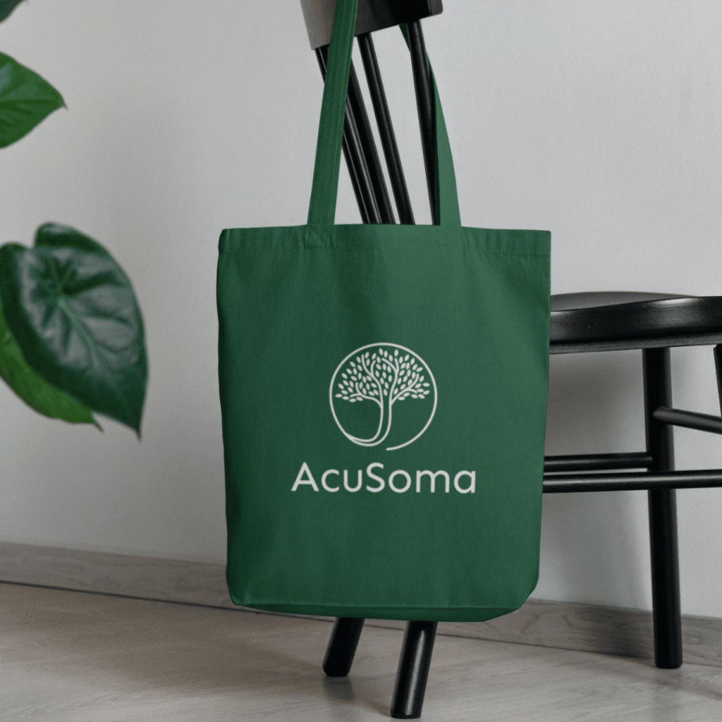 A green tote bag with the white AcuSoma logo and a stylized tree graphic, perfect for web designers or those seeking logo design services, hangs on the back of a black chair next to a plant.
