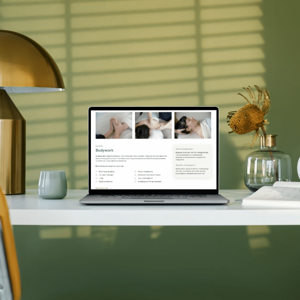 A laptop on a white desk displays a Moonlit Media custom website design for bodywork or massage therapy. The desk features a lamp, cup, vase with flowers, and sits against a green wall with light streaming in.