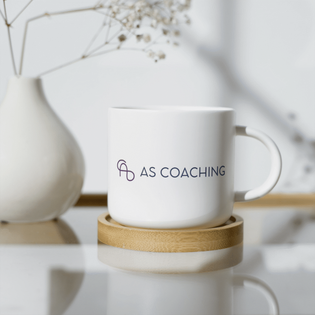 A white mug featuring AS COACHING and a minimalist logo—crafted with expert logo design services—rests on a wooden coaster. In the softly blurred background, a white vase holds delicate dried flowers.