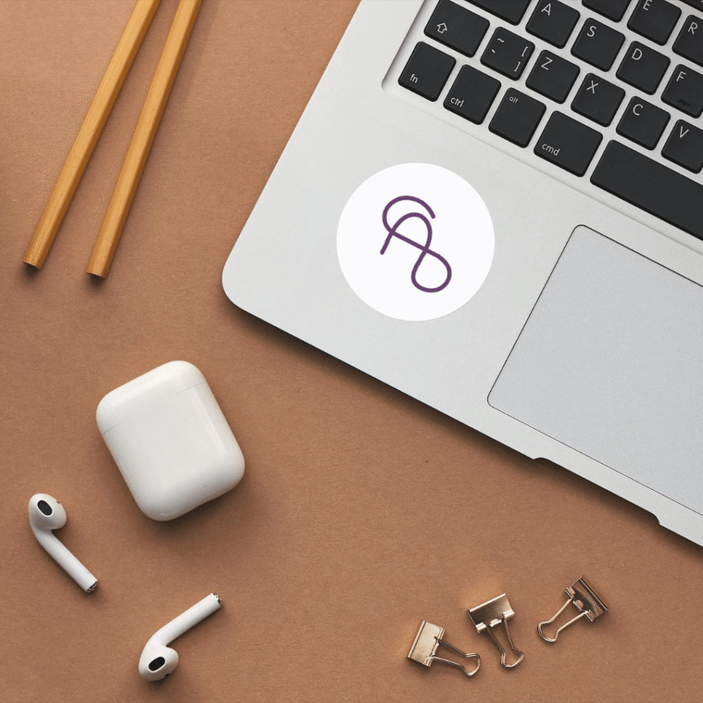 A laptop with a sticker, two brown pencils, wireless earbuds with an open charging case, and two metal binder clips are arranged on a brown surface—perfect for showcasing your logo design services.
