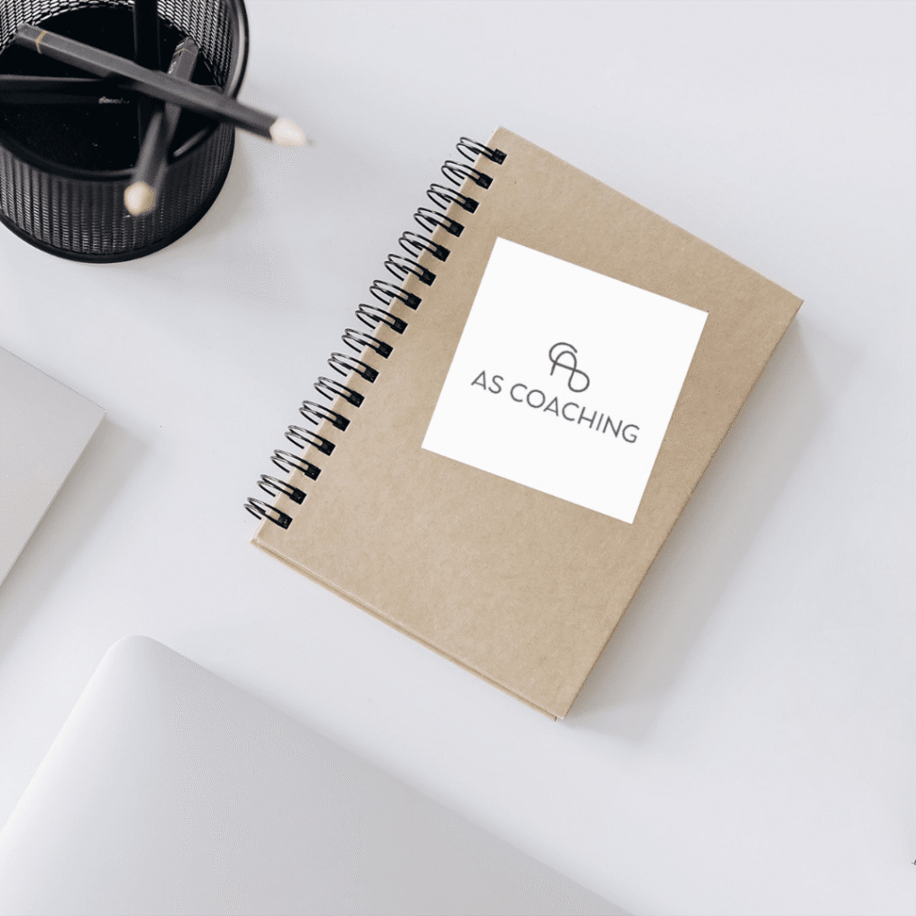 A spiral-bound notebook with a brown cover labeled AS COACHING lies on a white desk beside a pen holder with pencils, the corner of a laptop, and materials for custom logo design.