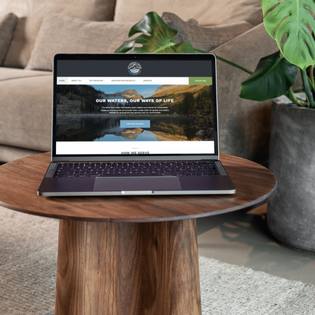 A laptop on a round wooden table displays a website by Moonlit Media, featuring a scenic lake and forest. The screen reads, “OUR WATERS, OUR WAYS OF LIFE.” A large potted plant and couch are in the background—true web design services at work.