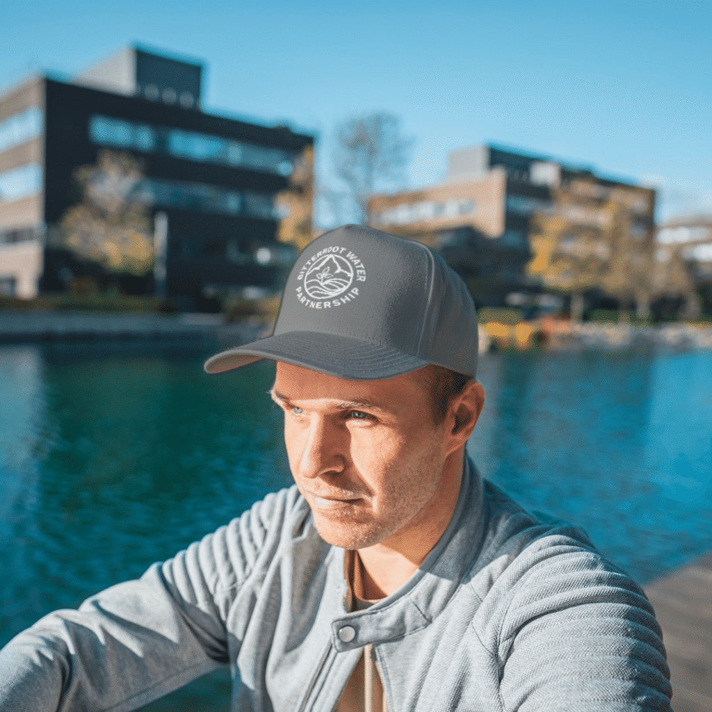 A man in a gray jacket and cap sits by a river, with modern office buildings—a hub for web design services—and trees in the background on a sunny day.