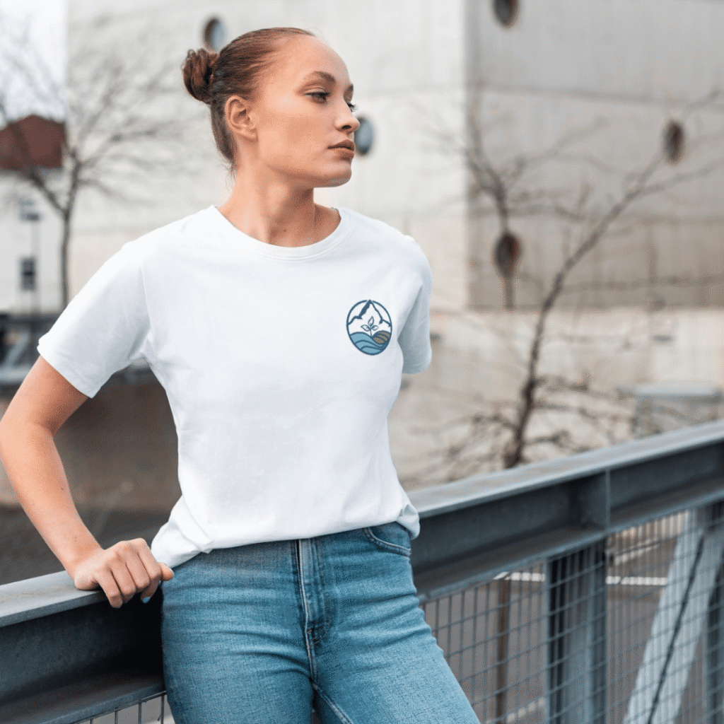 A woman with tied-back hair, wearing a white t-shirt featuring a custom logo design of a small mountain and wave and blue jeans, stands on an outdoor metal walkway, looking to the side. A modern building and bare trees are in the background.