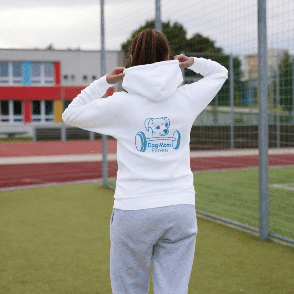 A person in a white Dog Mom Fitness hoodie and gray sweatpants stands on a sports field, facing away, near a soccer goal. The hoodie features a blue cartoon dog and barbell logo—perfect inspiration for any web designer or custom website design project.
