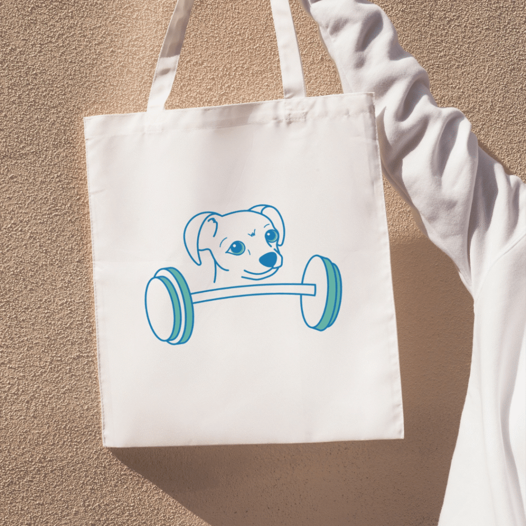 A person wearing a white long-sleeve shirt holds a white tote bag featuring a blue line drawing of a dog holding a barbell in its mouth—a playful touch perfect for any web designer or fan of Moonlit Media.