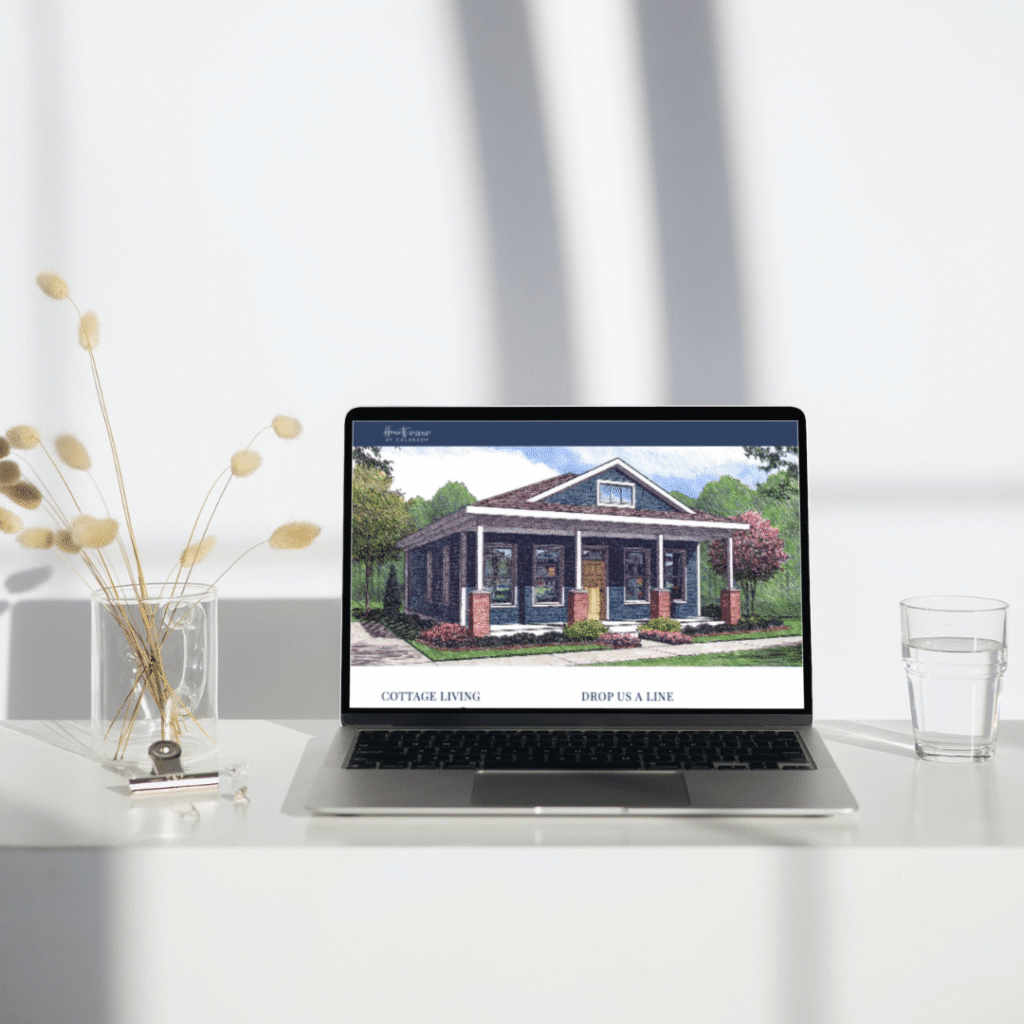 A laptop on a white desk displays a custom website design featuring an illustration of a cottage-style house; nearby are a glass of water and a vase with dried flowers, all lit by sunlight streaming from the left.