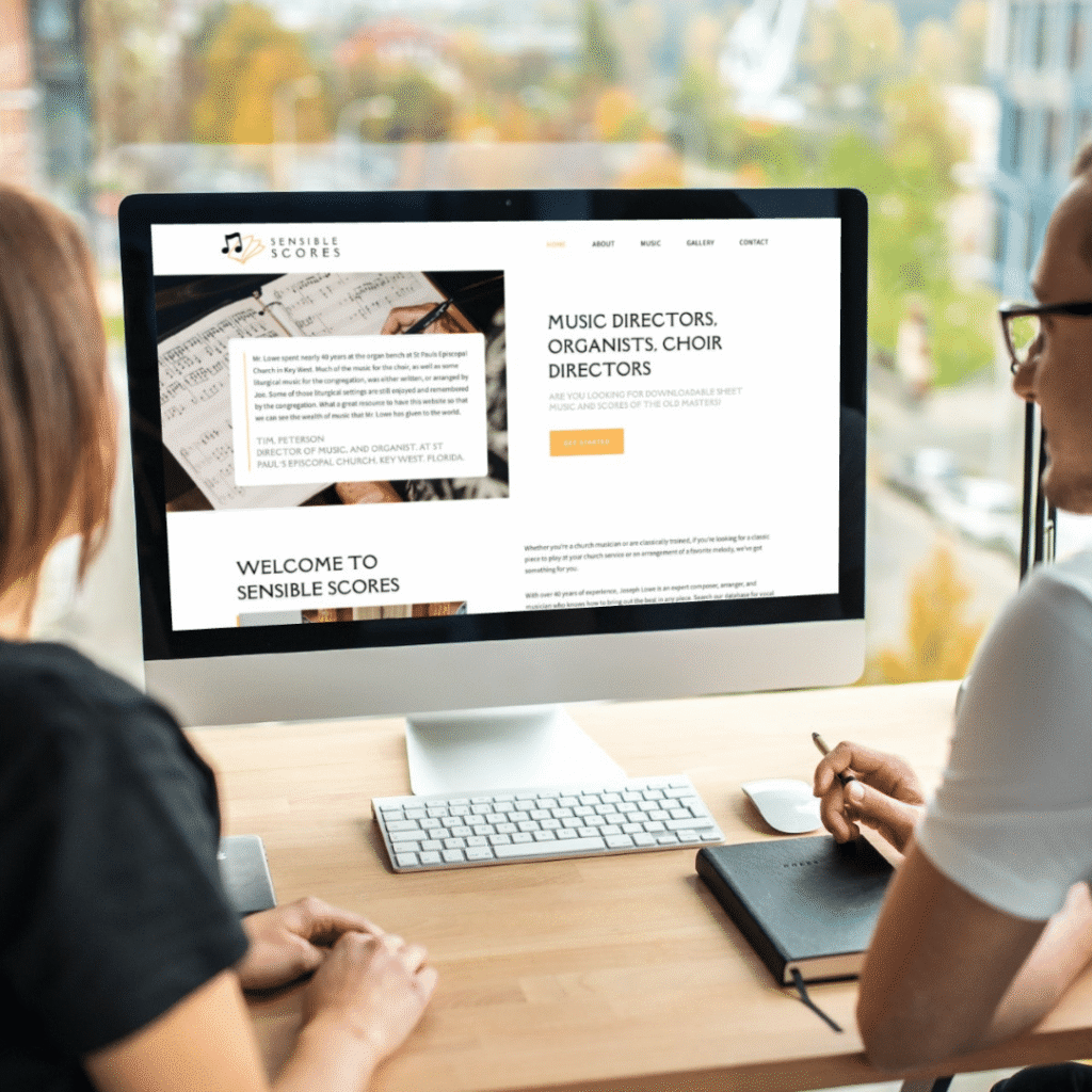 Two people sit at a desk looking at a computer screen displaying Sensible Scores, a custom website design featuring music sheets and information for music directors and choir directors. A notebook and pen are on the desk.