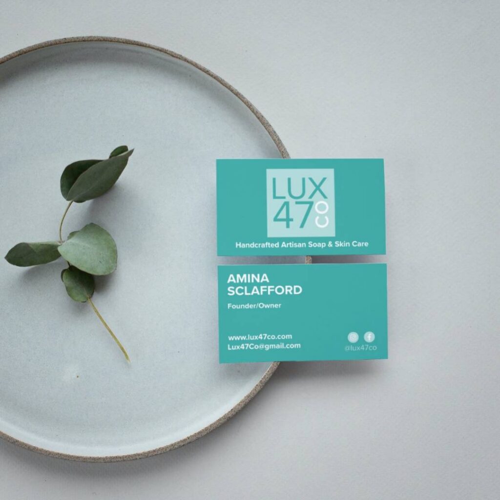 | Moonlit Media LLC Two turquoise business cards for LUX 47 Co rest on a light gray plate next to a sprig of eucalyptus. The cards display the company name, tagline, and details for Amina Sclafford, Founder/Owner, specializing in custom logo design and web design services.